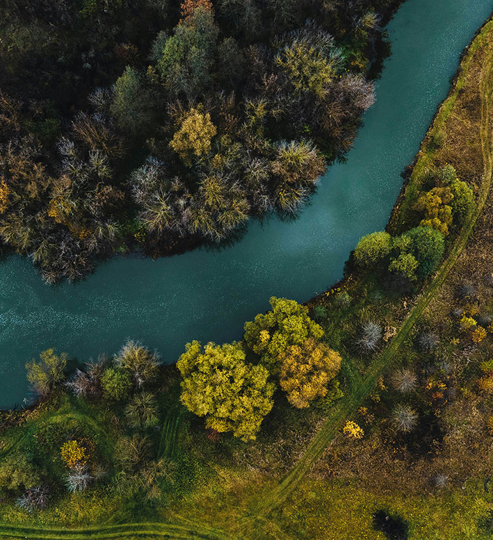 An aerial view of a winding, dark blue-green river flowing through a landscape of trees with autumn foliage.