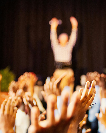 Audience with their hands raised enthusiastically in a crowd mirroring the speaker on stage.