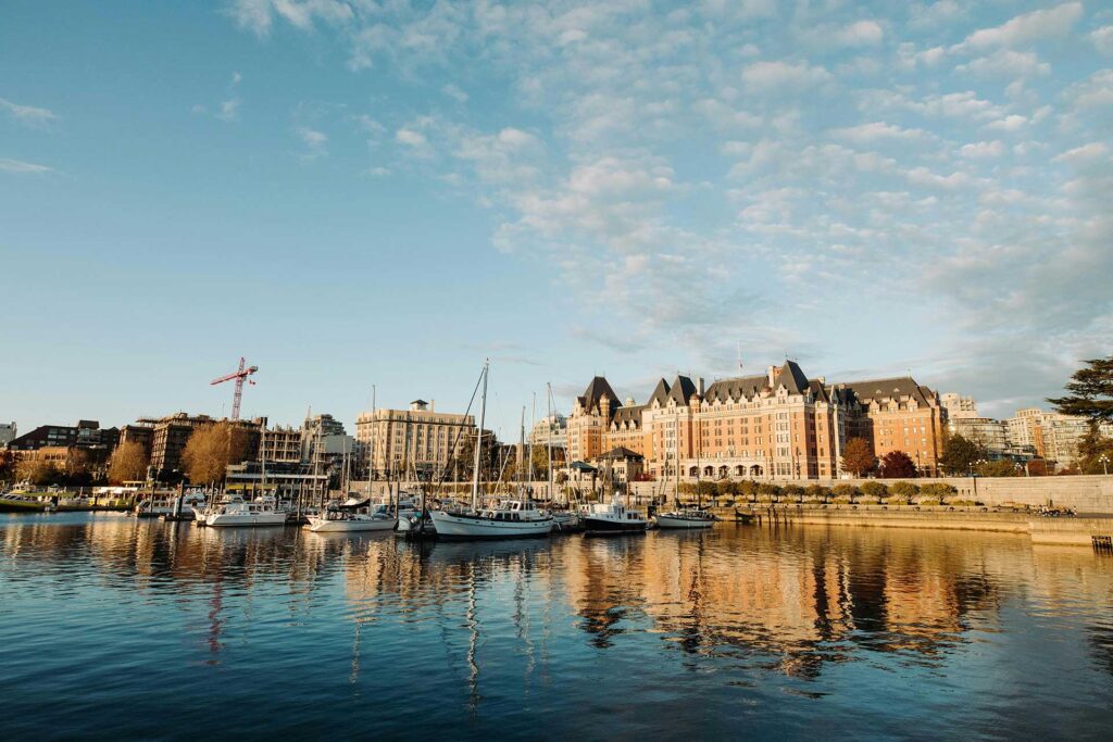 View of Victoria, BC harbour and the Empress Hotel from the water with boats in the marina under a blue sky.